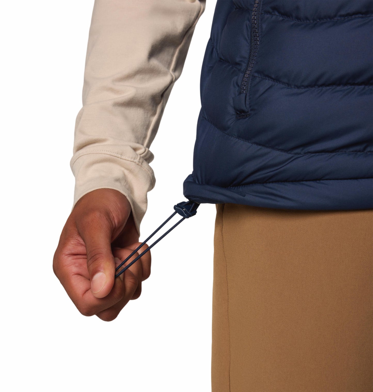 A man wearing a blue quilted vest over a beige long-sleeve shirt and brown pants. The vest has a zipper front and the man has his hands in the zippered hand pockets.