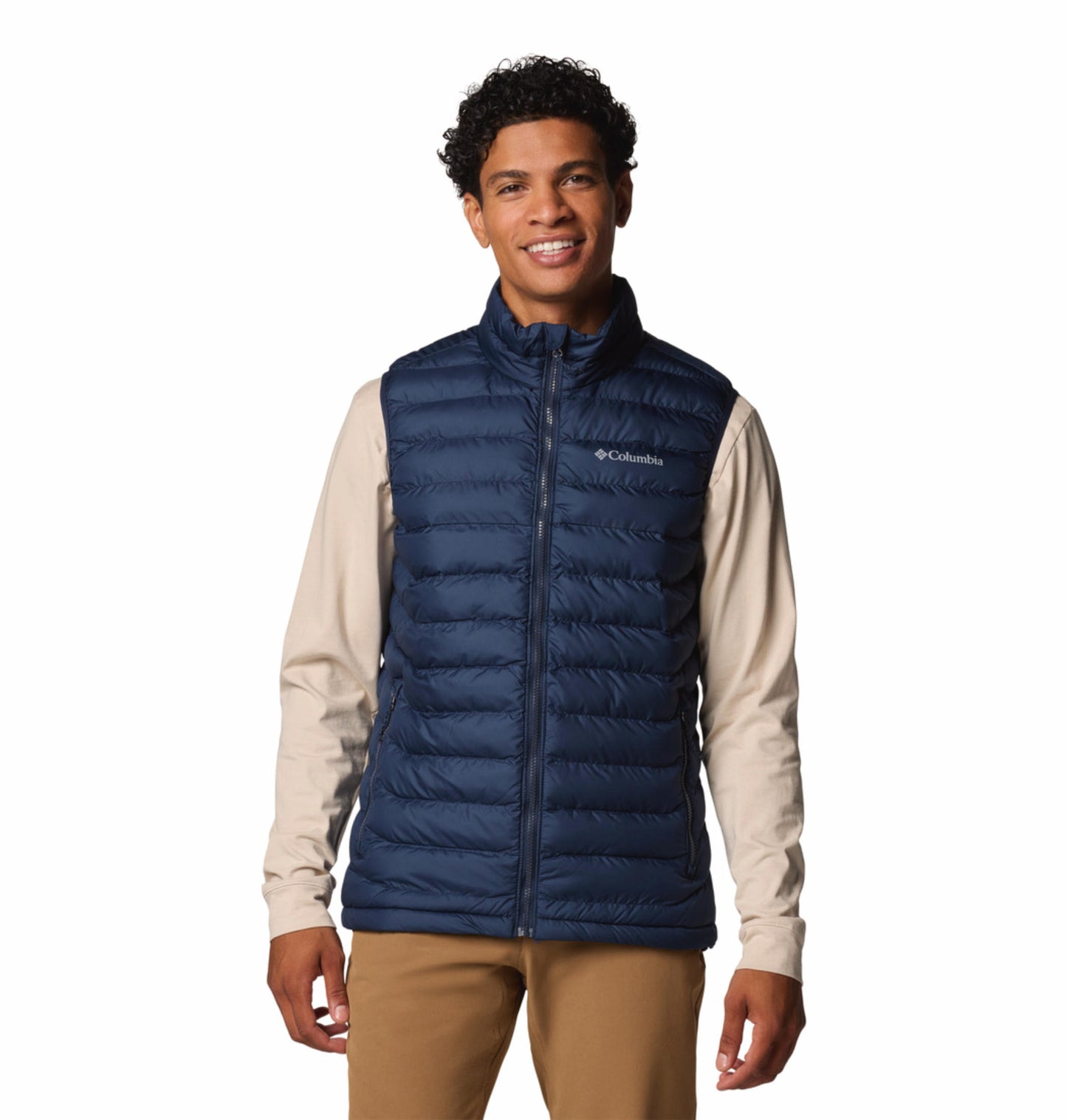 A man wearing a blue quilted vest over a beige long-sleeve shirt and brown pants. The vest has a zipper front and the man has his hands in the zippered hand pockets.