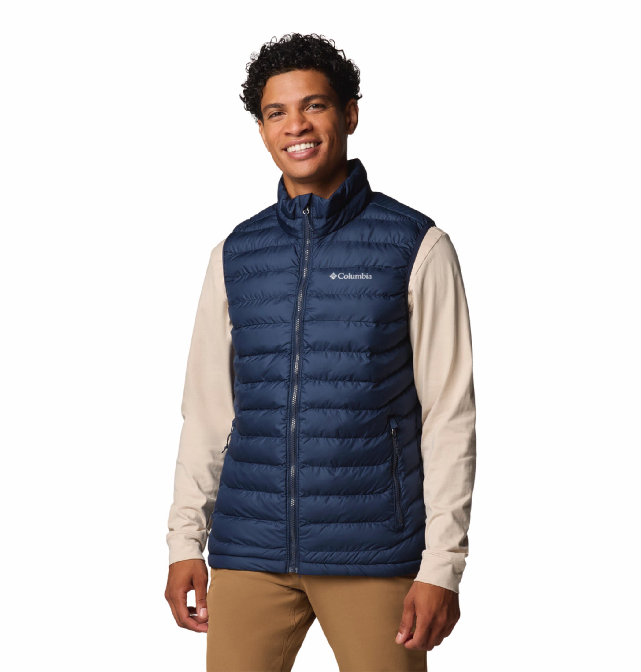 A man wearing a blue quilted vest over a beige long-sleeve shirt and brown pants. The vest has a zipper front and the man has his hands in the zippered hand pockets.
