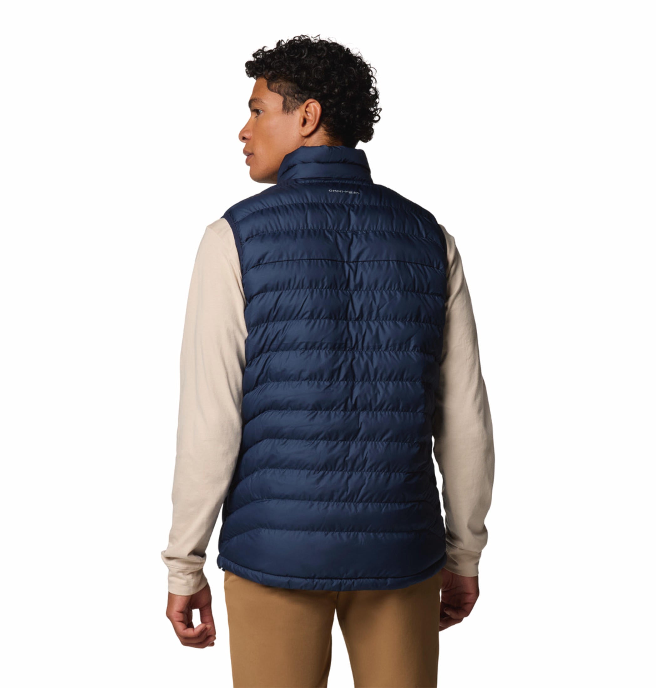 A man wearing a blue quilted vest over a beige long-sleeve shirt and brown pants. The vest has a zipper front and the man has his hands in the zippered hand pockets.
