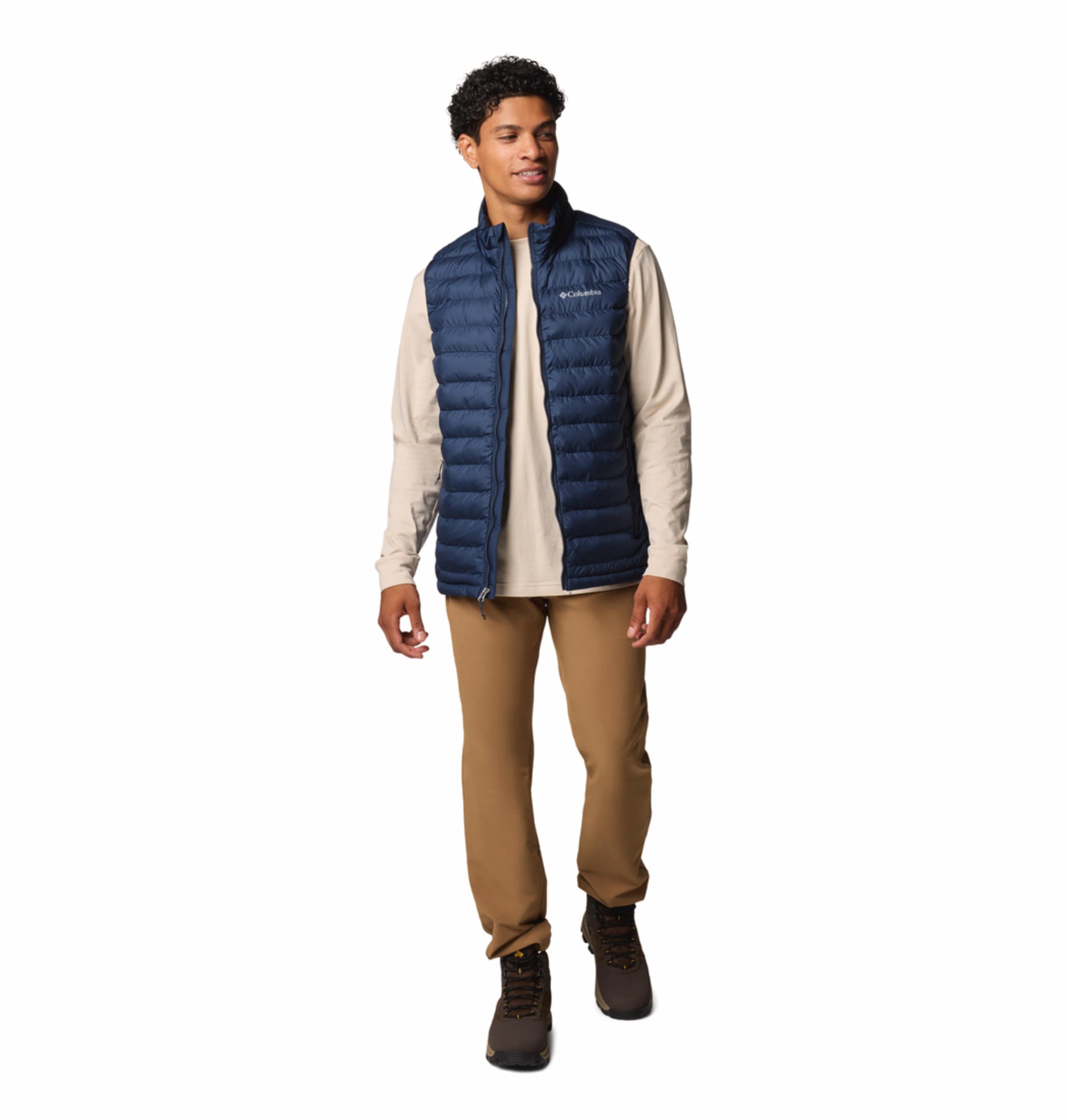 A man wearing a blue quilted vest over a beige long-sleeve shirt and brown pants. The vest has a zipper front and the man has his hands in the zippered hand pockets.