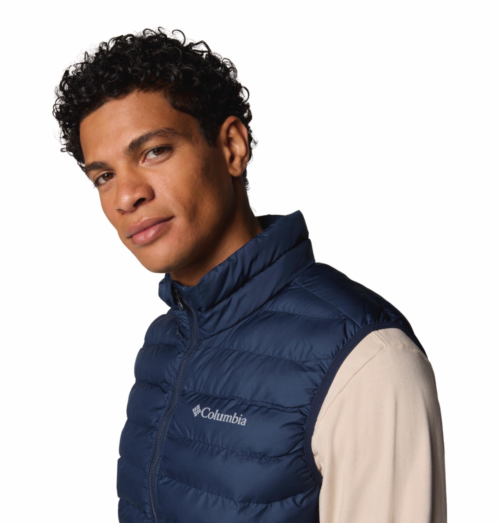 A man wearing a blue quilted vest over a beige long-sleeve shirt and brown pants. The vest has a zipper front and the man has his hands in the zippered hand pockets.