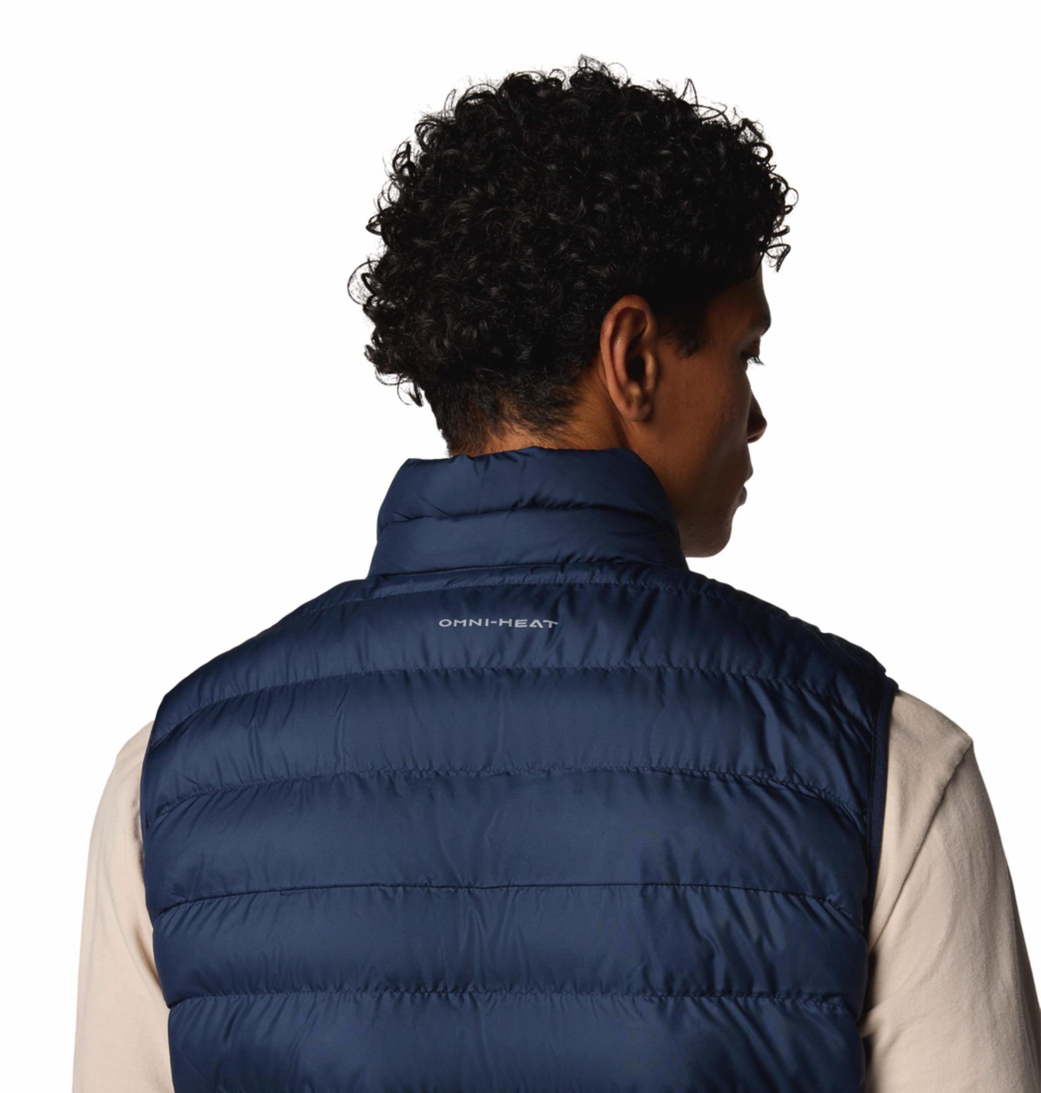 A man wearing a blue quilted vest over a beige long-sleeve shirt and brown pants. The vest has a zipper front and the man has his hands in the zippered hand pockets.